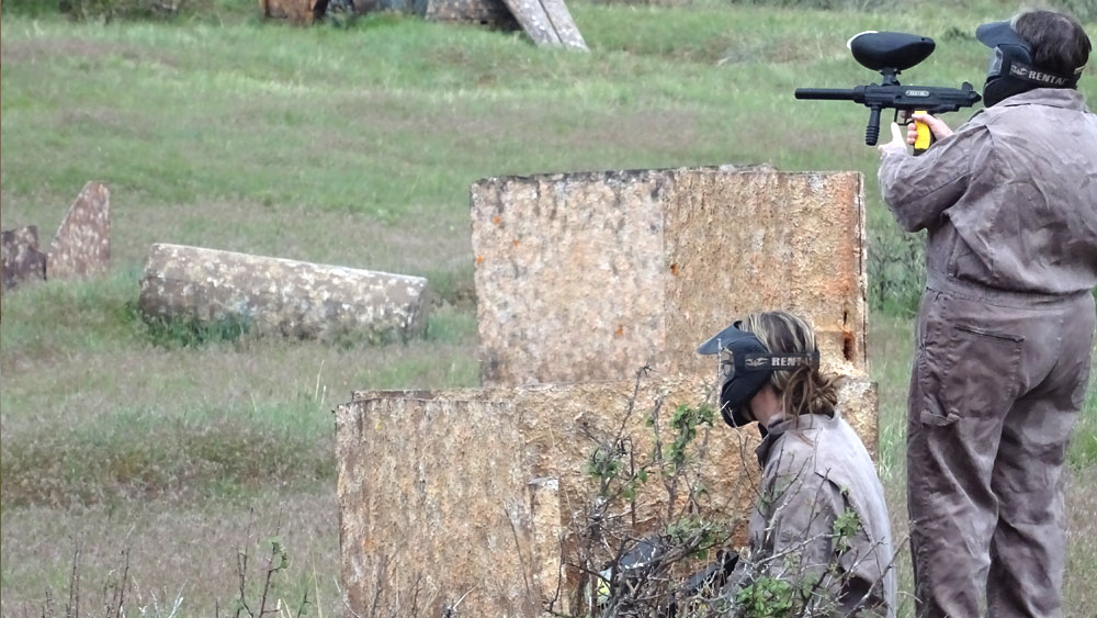 Two players in paintball gear take cover behind barriers in a grassy field.